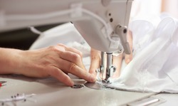 Close up view of sewing process. Female hands stitching white fabric on professional manufacturing machine at workplace. Seamstress hands holding textile for dress production. Light blurred background