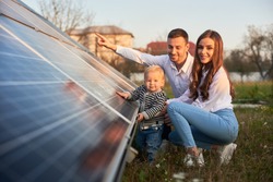 A young family of three is crouching near a photovoltaic solar panel, smiling and looking at the camera, concept of bright future