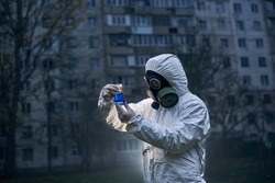 Ecologist wearing white coverall and gas mask holding glass flask with samples, observing reaction on the light, high building on background