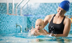 Young attentive mother is helping her little son swim in turquoise water during swimming class for infants, baby is splashing around in water. Concept of active family leisure