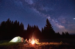 Night camping near bright fire in spruce forest under starry magical sky with milky way. Group of four friends sitting together around campfire, enjoying fresh air near tent. Tourism, camping concept.