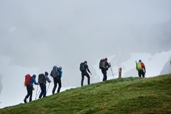 Group of male tourists with backpacks climbing grassy hill. Brave alpinists walking uphill in mountains with foggy cliff on background. Concept of hiking, travelling, mountaineering and backpacking.