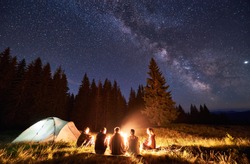 Night summer camping in the mountains, spruce forest on background, sky with stars and milky way. Back view group of five tourists having a rest together around campfire, enjoying fresh air near tent.
