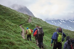 Back view of travelers with backpacks using trekking poles while climbing the grassy hill. Group of male hikers walking on path and heading to foggy mountain. Concept of hiking, climbing and alpinism.