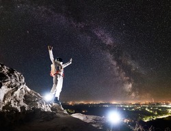 Astronaut raising arms while standing on rocky mountain with fantastic starry sky with Milky way on background. Spaceman wearing white space suit and helmet. Concept of space travel