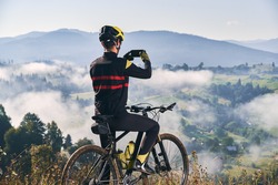 Back view of man sitting on bicycle and talking mountain photo with smartphone. Male bicyclist holding cellphone and taking picture of beautiful misty hills. Concept of sport, cycling and photography.