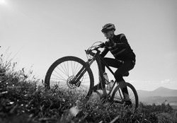 Smiling man in safety helmet and glasses cycling uphill on sunny day. Male bicyclist in cycling suit climbing uphill on mountain bike. Concept of sport and active leisure. Monochrome image.