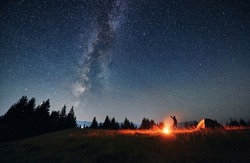 Man hiker standing on grassy hill near campfire and pointing finger at beautiful night starry sky with Milky Way under mountain valley. Concept of hiking, night camping and astrology.