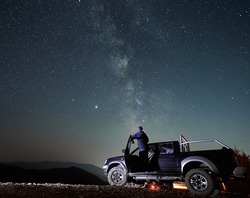 Rear view of young man who enjoying magic Milky Way and many stars in night sky looking out of the cabin black car. Night riding on stony mountain roads by big car.