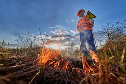 Firefighter ecologist working in field with wildfire in evening. Man in orange vest and helmet near burning grass with smoke, holding warning sign with exclamation mark. Natural disaster concept.