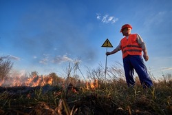 Fireman ecologist extinguishing fire in field in evening. Man in orange vest and helmet near burning grass with smoke, holding warning sign with exclamation mark. Natural disaster concept.