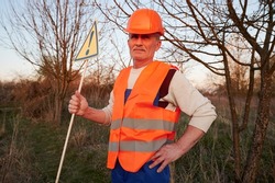 Fireman ecologist extinguishing fire in field in evening. Man in orange work vest and safety helmet near burning grass with smoke, holding warning sign with exclamation mark. Natural disaster concept.