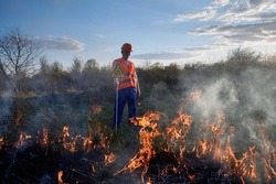 Fireman ecologist working in field with wildfire in evening. Man in orange vest and helmet near burning grass with smoke, holding warning sign with skull and crossbones. Natural disaster concept.