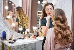 Female stylist doing professional makeup for client in beauty salon. Young woman sitting at dressing table and looking in the mirror while makeup artist applying concealer with cosmetic brush.