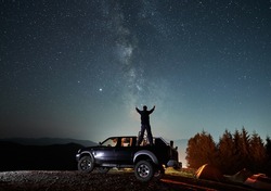 Rear view of boy standing at night on surface of body car parked on dirt mountain road. Boy stretched his arms out to sides as if reaching them to starry sky. Nearby tourist camp with three tents.