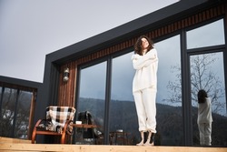 Curly young woman resting on terrace of modern barn house in the mountains. Happy female tourist enjoying in new cottage in winter.