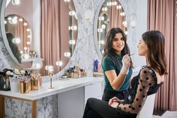 Portrait of makeup artist smiling to camera while doing professional makeup in visage studio. Woman sitting at dressing table while female beauty specialist applying lipstick with cosmetic brush.