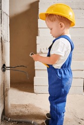 Adorable child construction worker using wire stripper cutter tool while installing electric cables and socket in wall. Kid in safety helmet mounting electrical wiring in apartment under renovation.