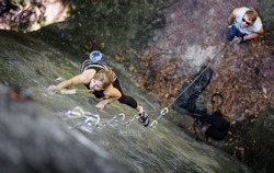 Athletic young woman rock climbing with carbines and rope on summer day. Man standing on the ground insuring the climber
