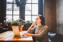 Theme is small business. A young freelance woman working behind a laptop computer in a coffee shop decorated with Christmas decor and talking on the phone. Dressed in a gray sweater and glasses.