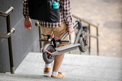 Man Carrying folded E-Scooter in hand on stairs near building. Moving by eco urban transport, modern city concept. Ecological technological lifestyle. Person Carry Electric Scooter In Folded Position.