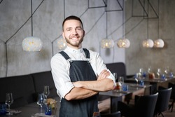 A young, smiling waiter in a restaurant, standing next to the tables with a glass of wine. He wears an apron, looks confidently, folded his arms over his chest