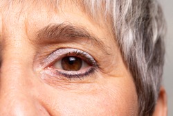 A closeup view on the eye of an old-aged woman waring blue eye shadow and eyeliner. Lady with brown eyes and grey hair with wrinkled face.