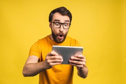 Amazed happy bearded man using digital tablet looking shocked about social media news, astonished man shopper consumer surprised excited by online win isolated over yellow background.