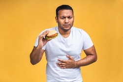 Young african american indian black man eating hamburger isolated over yellow background. Unhealthy food, stomach pain.