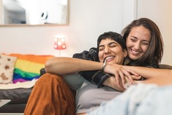 Authentic shot of happy married homosexual female gay couple laughing and embracing on the sofa with rainbow pride flag on background - lesbian couple at home enjoying life together