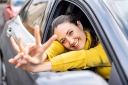 Happy woman portrait in a car in the city, looking out of the window, showing victory peace sign with fingers and smiling at camera - Travel and vacations concepts