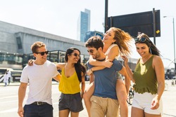 Multiracial group of friends together in the city. Multi ethnic group in London, walking on pavement on a sunny day. Man giving a piggyback ride to a girl. Friendship and lifestyle concepts
