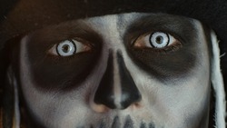 Close-up shot of man face in skeleton Halloween cosplay costume. Guy in creepy skull makeup opening eyes with white pupil, trying to scare. Day of The Dead. Isolated on black background