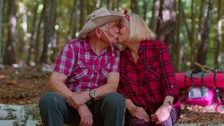 Old elderly married couple resting in forest. Grandmother grandfather senior tourists sitting on tree and hugging, kissing. Mature man woman embracing in wood. Active hiking and tourism outdoors