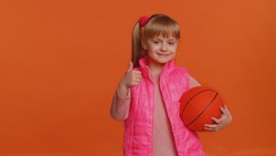 Girl sportsman basketball fan holding ball looking at camera, training dribbling. Workout sport motivation lifestyle concept. Young little blonde child kid isolated alone on orange studio background