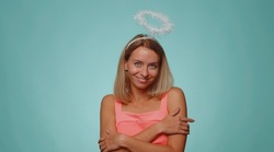 Portrait of smiling shy angelic blonde young woman with angel halo nimb over head flirting, looking at camera, positive emotions, celebrating holiday. Pretty girl isolated on studio blue background