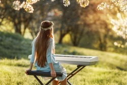 Girl playing a synthesizer piano in nature at sunset. A woman is looking at the sun during sunset. An electric piano stands in a field in a village. Spring day in the countryside. 