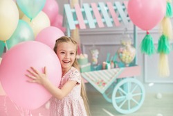 A portrait of a beautiful little girl smiles and holds in a hands a big color balloon in the studio with many balloons and a toy candy shop