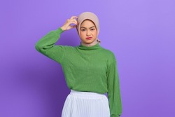 Beautiful young Asian woman in green sweater confused about a question, thinking with hand on head isolated over purple background