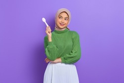 Hungry beautiful Asian woman in green sweater holding spoon think of tasty food isolated over purple background