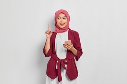 Excited beautiful Asian woman in casual shirt and hijab holding mobile phone and pointing finger up, creating genius solutions isolated over white background. People religious lifestyle concept