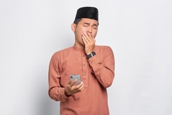 Confused young Asian Muslim man holding mobile phone and feeling sleepy isolated over white background