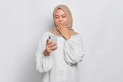 Confused young Asian Muslim woman holding mobile phone and feeling sleepy isolated over white background