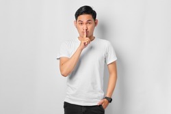 Portrait of serious young Asian man showing a sign of silence gesture putting fingers in mouth isolated on white background