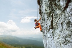 side view of young slim woman rock climber in bright orange pants climbing on the cliff against a blue sky. girl climbs on a vertical flat rocky wall and making hard move. Copy space