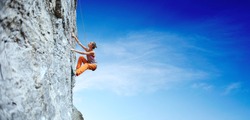 side view of young slim woman rock climber in bright orange pants climbing on the cliff. a woman climbs on a vertical rock wall on the blue sky background