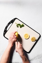  Shot of hands holding a knife and cuting a lemon on a white cutting board with black inserts at the edges. There are some green herbs and lemon slices on the cutting board. 