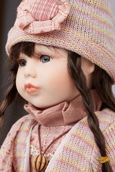Medium close-up shot of a porcelain doll in retro clothes: in a turtleneck dress, a jacket, a hat with a flower and a pendant on a cord. The doll with brown pigtails is located on grey background. 