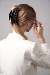 A back turned blonde girl with golden earrings and rings is wearing a white blouse. The lady's hair is fixed with semicircular black hair clip. The woman is posing on the gray backdrop.