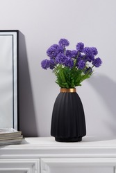 Subject shot of a graceful bouquet of tender violet asters in a black ceramic vase. The vase with a bunch of flowers is located on the white table on the white background. 
